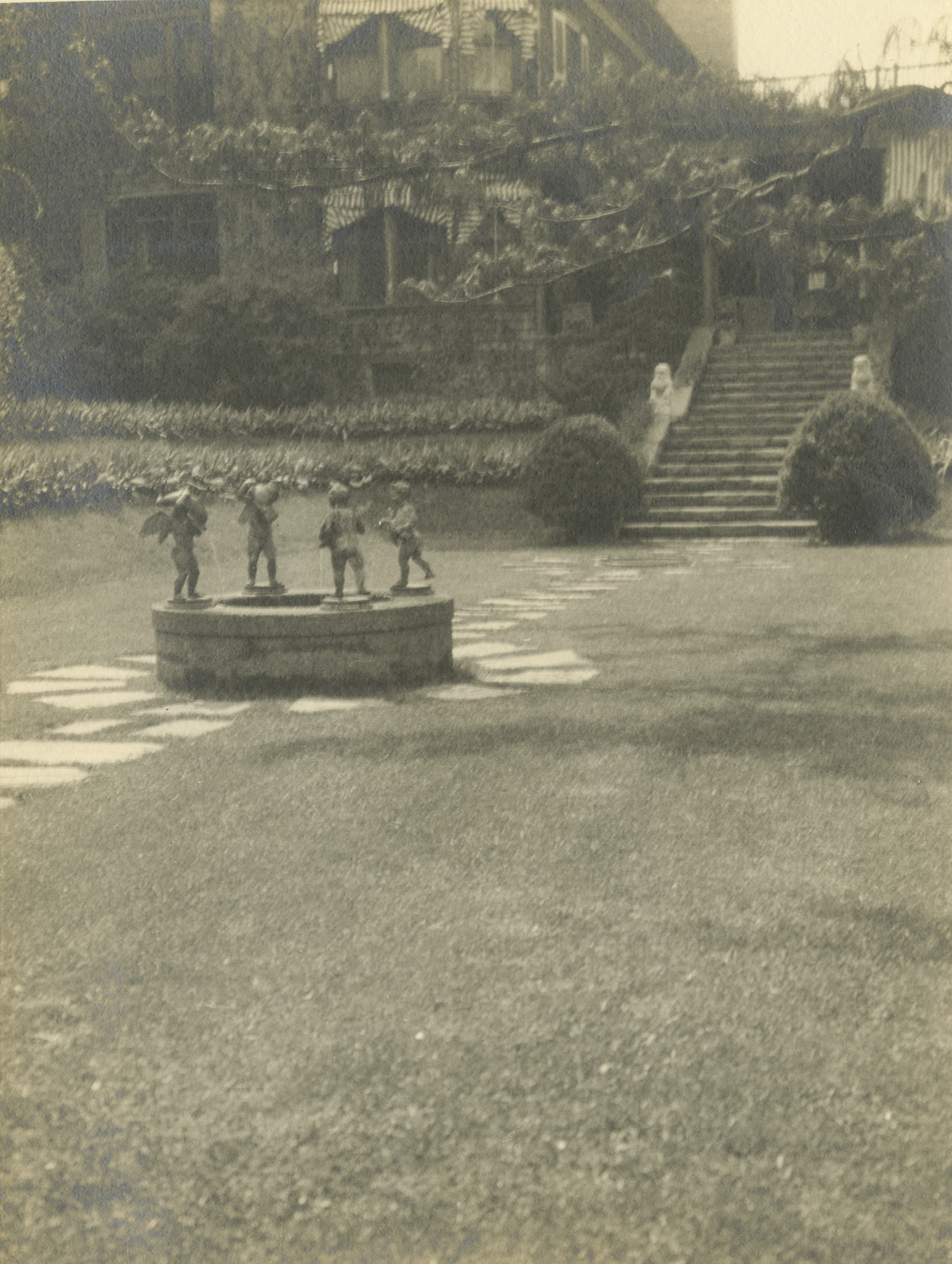 An image of a Taft garden. In foreground a fountain with four angles can be seen. Behind are steps which have two Chinese stone lions sitting on either side of the stone wall